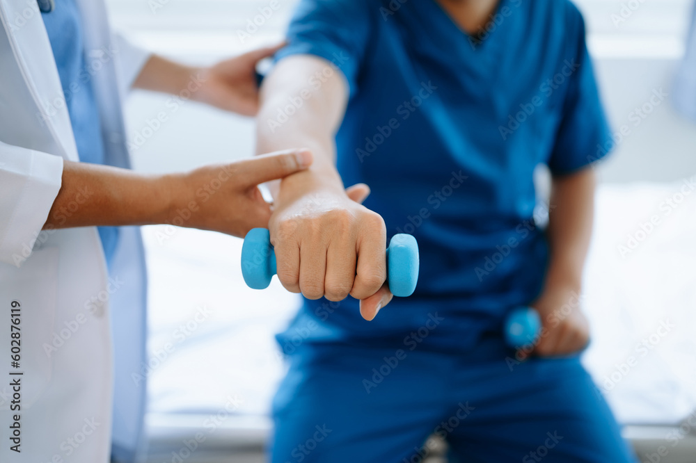 Asian physiotherapist helping male patient stretching arm during exercise correct with dumbbell in hand during training hand in hospital office. Asian physiotherapist helping male patient stretching arm during exercise correct with dumbbell in hand during training hand in hospital office.