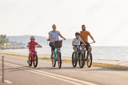 Two kids, with helmets on their heads, and smiling parents riding bikes on a family-friendly cycle route along a sea coastline, front view.