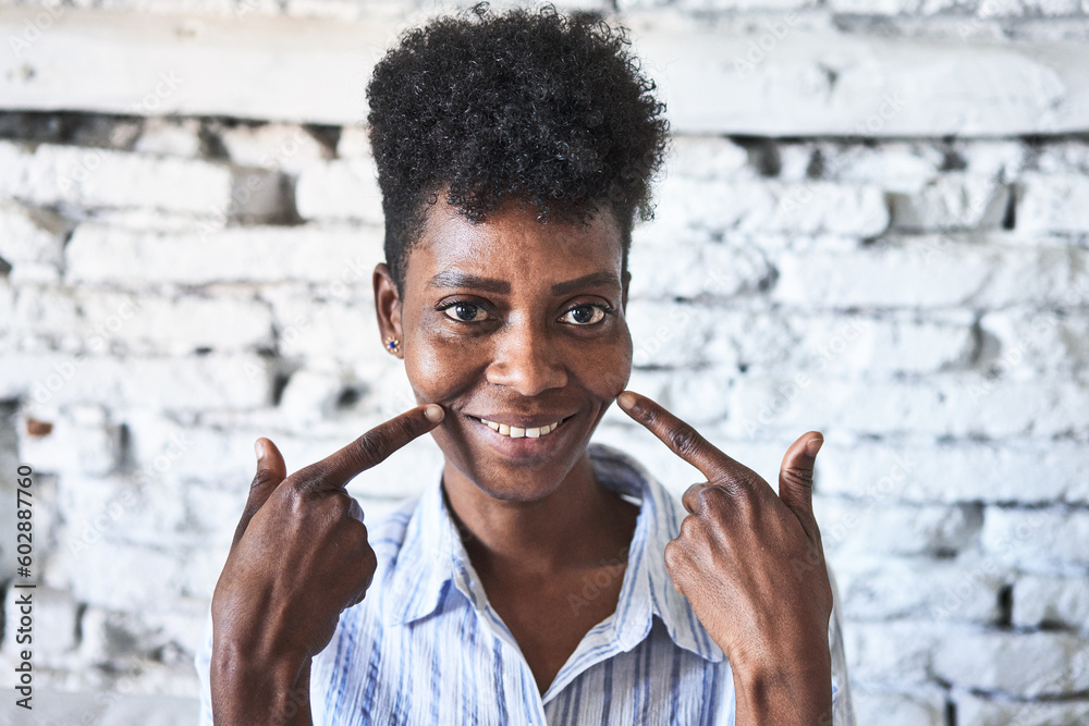 Obraz premium Radiant middle-aged African woman smiles in her living room with white brick wall and natural light.