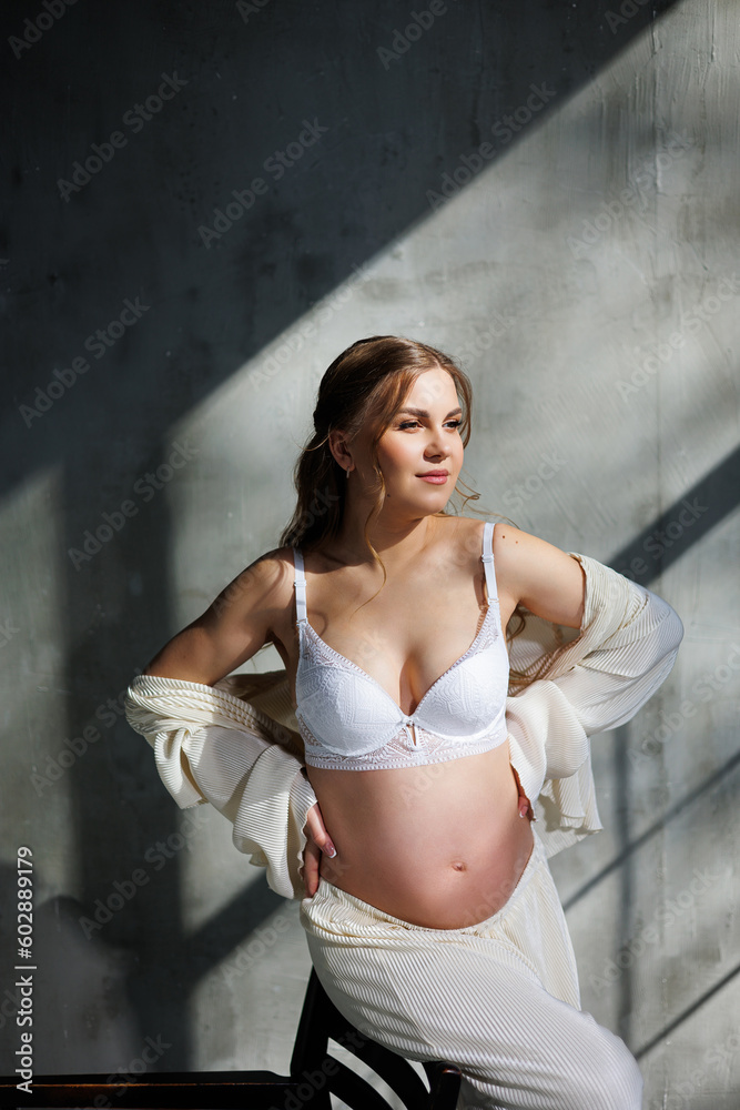 A stylish young pregnant woman in a white suit is sitting on a chair on a gray background. Pregnant woman in white underwear. A stylish young pregnant woman in a white suit is sitting on a chair on a gray background. Pregnant woman in white underwear.