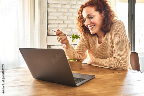 Redhead woman enjoys salad while watching streaming content on laptop, relaxed.