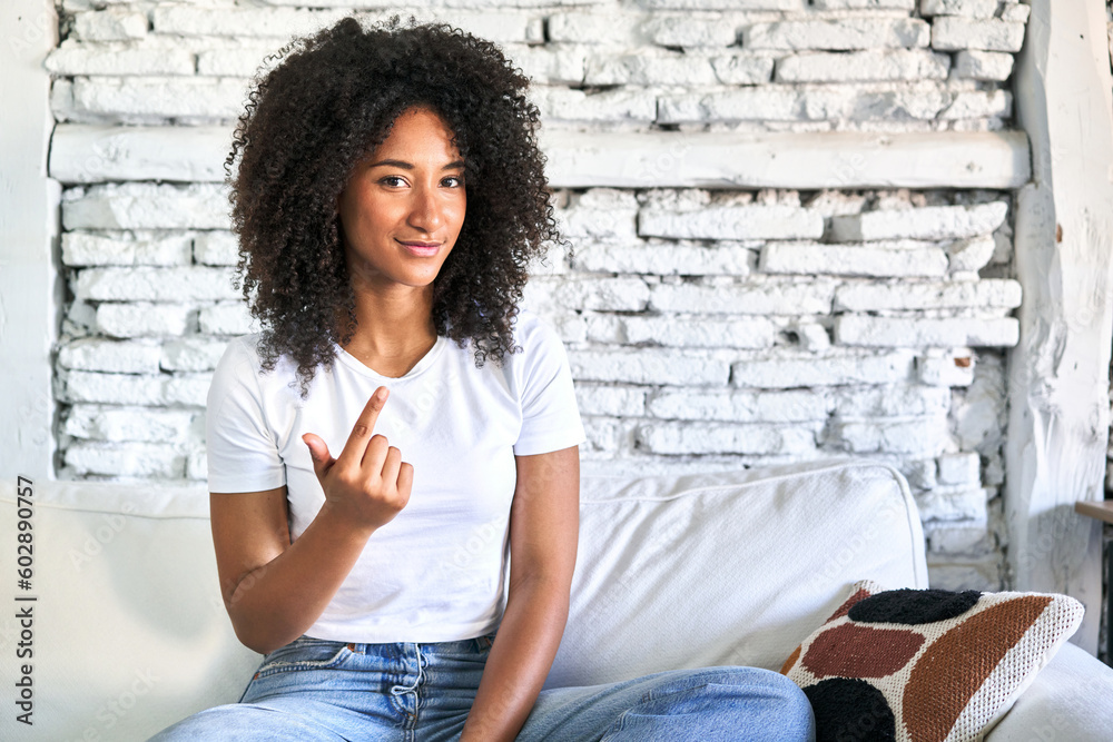A young african american woman sitting on her sofa pointing with finger at you as if inviting come closer.