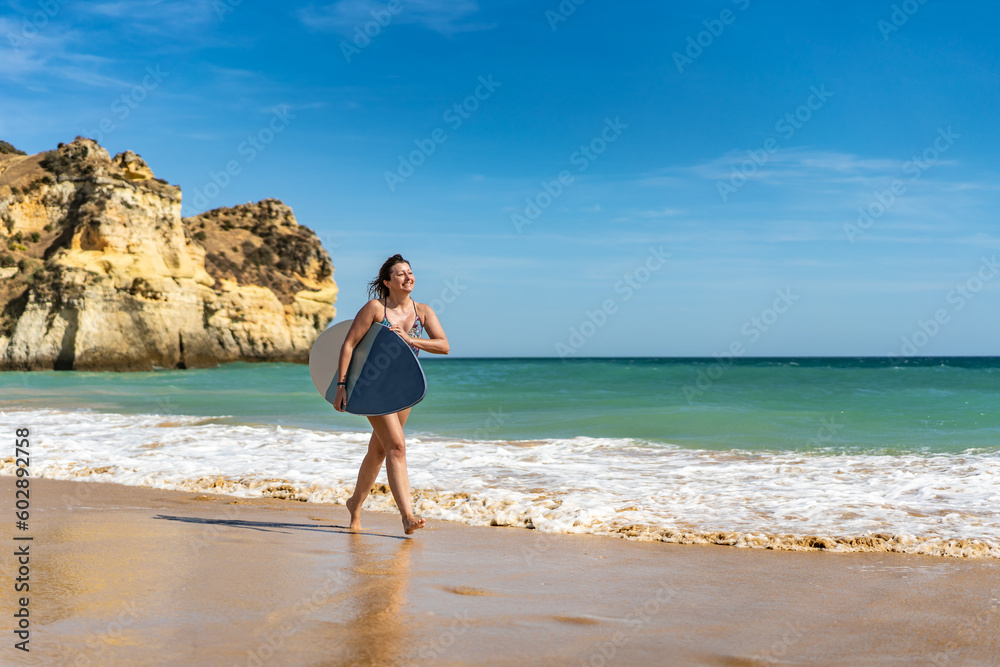 Beautiful woman holding  skimboard walking on sunny beach 
