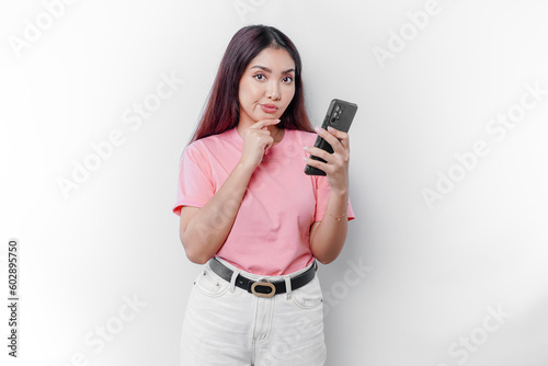 A thoughtful young woman wearing pink t-shirt, holding her chin and her phone isolated by white background