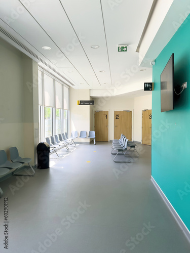 A hospital waiting room with empty chairs, numbered cabins and a large plasma TV. An architecture of stark flooring contrasted against the clinical interior.