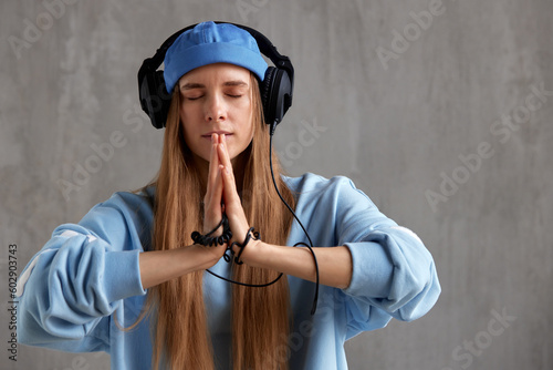 A young charming red-haired woman in a blue sweater and cap with DJ headphones on her head folded her hands in a prayer pose. Music and meditation.