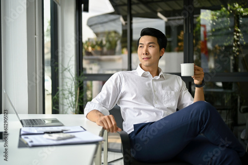 Confident Asian businessman sits at his desk in modern office with a coffee cup