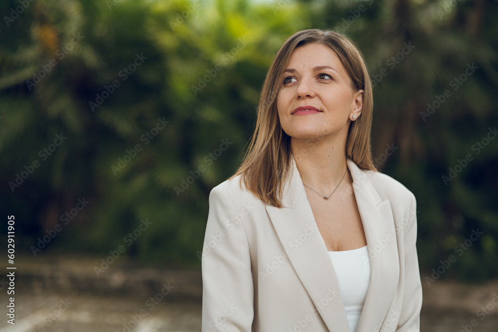 Dreamy businesswoman in jacket standing against greenery Dreamy businesswoman in jacket standing against greenery