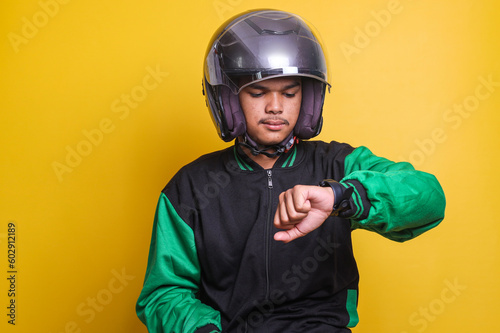 Asian online taxi driver wearing green jacket and helmet checking the time on wrist watch isolated over yellow background