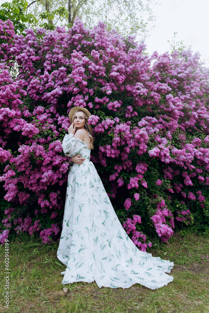 Beautiful, stylish, cute blonde in a white dress with a green pattern as a model for a photo shoot in lilac. They made luxurious portraits of her in the green garden of the park.