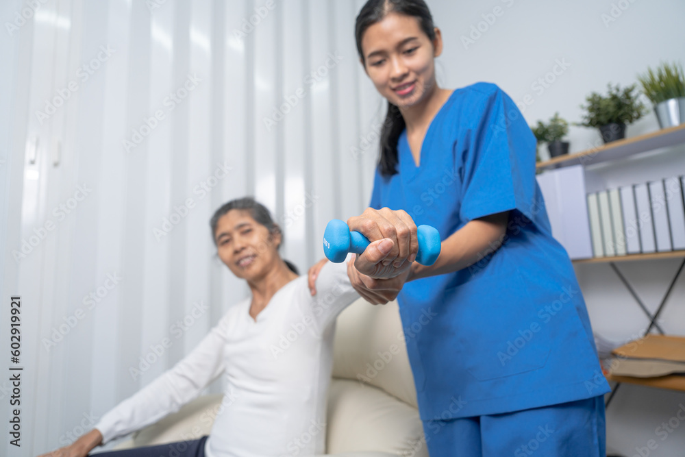 Fototapeta premium Young pretty asian physiotherapist helping senior female patient holding dumbbell in physical therapy session, concept of World Confederation for Physical Therapy.