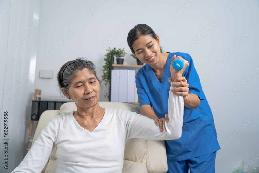 Fototapeta premium Young pretty asian physiotherapist helping senior female patient holding dumbbell in physical therapy session, concept of World Confederation for Physical Therapy.