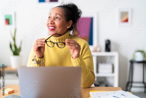Pensive elderly Ethiopian female employee with eyeglasses looking away and smiling while analyzing business plan and working remotely with portable computer photo