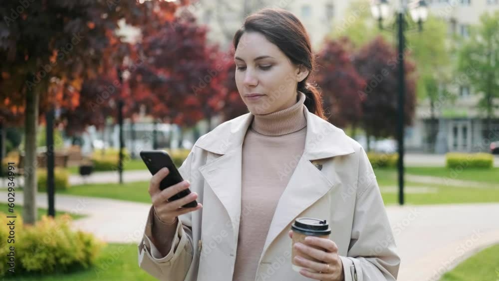 An attractive young woman with her hair pulled back in a ponytail is standing on the street and using a smartphone in a park against a background of green bushes. Digital technologies, mobile Internet