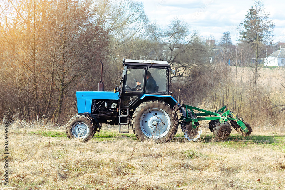 Obraz premium Blue small modern tractor machinery plowing agricultural field meadow at farm on sunny day. Farmer cultivating and make soil tillage before seeding plants and crops, nature countryside rural scene