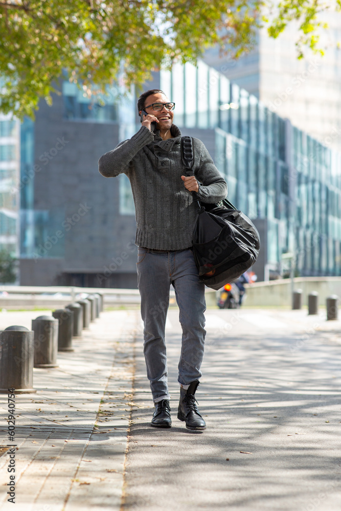 Cool young guy talking on cell phone carrying bag in town