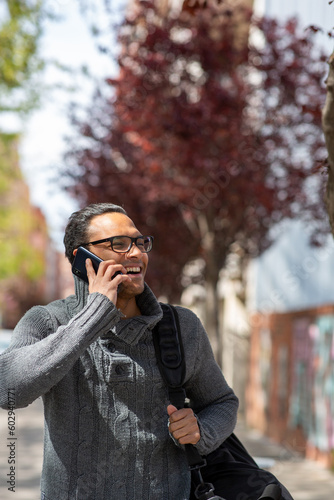 Smiling young african guy with bag walking outside and talking on mobile phone