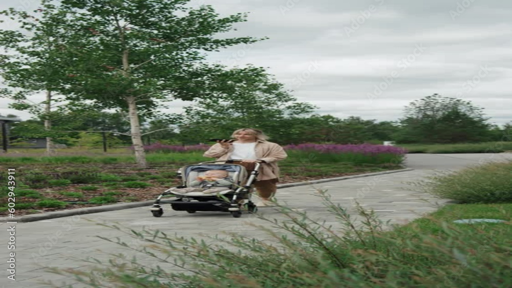 Vertical long shot of young mother recording voice message on smartphone while walking with her baby in stroller Vertical long shot of young mother recording voice message on smartphone while walking with her baby in stroller