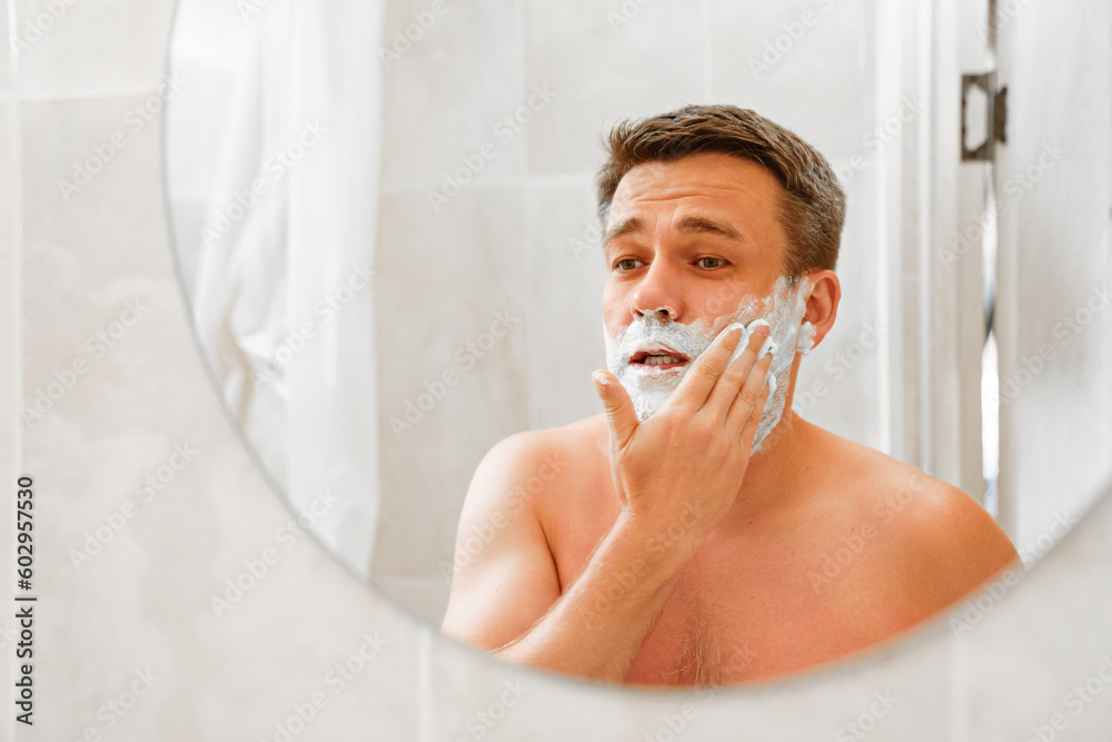a man applies shaving foam on his face and looks in a round mirror. The