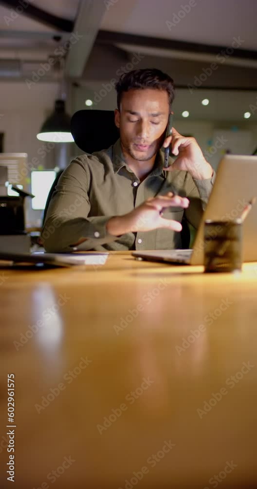 Vertical video of latino businessman using laptop and talking on smartphone at office, slow motion
