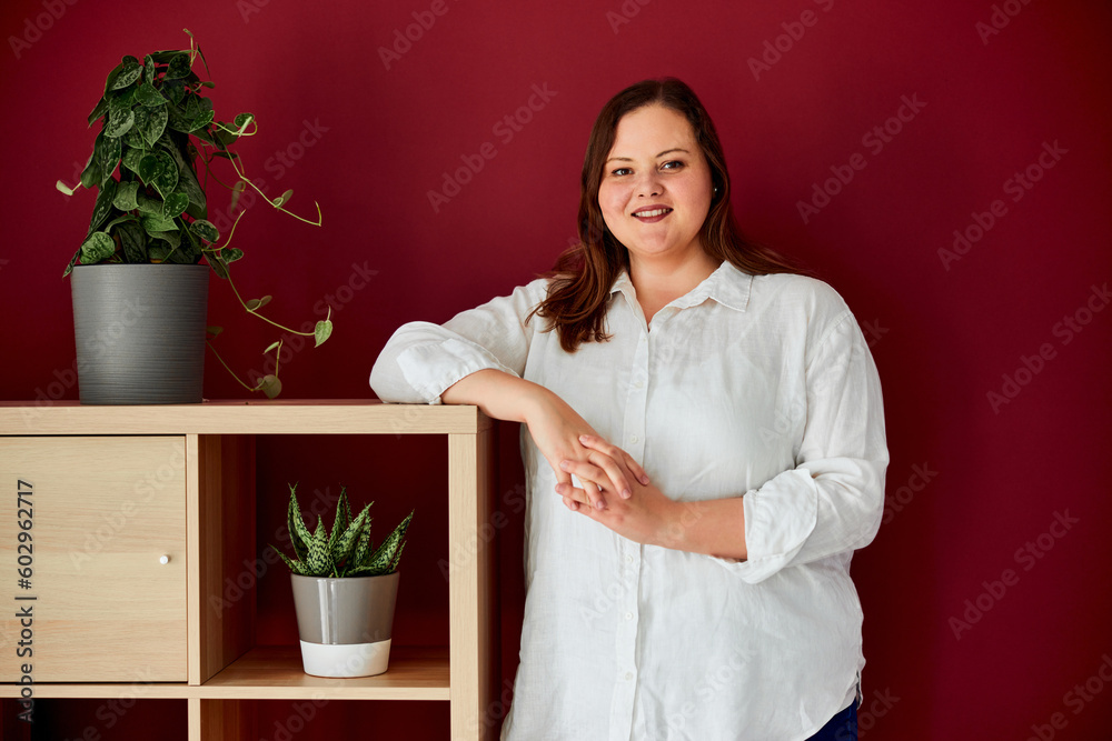 Portrait of a happy plus-size woman standing in front of the red wall next to the wooden shelf. Portrait of a happy plus-size woman standing in front of the red wall next to the wooden shelf.
