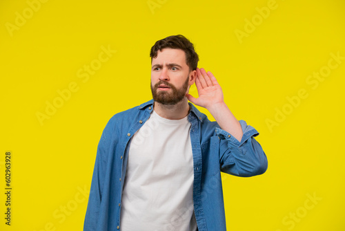 Quadro su tela Young, serious, blond man in a blue shirt on a yellow background, holding an ear to listen better, what you're saying