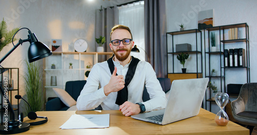Likable positive satisfied confident young bearded man in office clothes poses on camera with thumbs up on the home office background,front view © serg