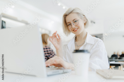 Young beautiful joyful blonde woman influencer blogger smiling while working with laptop in cafe with coffee  white light