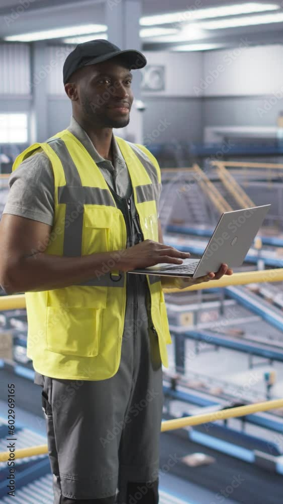 Vertical Screen: Black Man Standing on a Metal Platform, Using a Laptop Computer at a Post Office Logistics Center. African Specialist Working at a Sorting Facility, Configuring the Automation Process Vertical Screen: Black Man Standing on a Metal Platform, Using a Laptop Computer at a Post Office Logistics Center. African Specialist Working at a Sorting Facility, Configuring the Automation Process