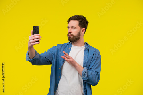 Happy young man video calling, talking online with mobile phone, saying hello to smartphone camera and waving hand friendly, standing over yellow background © kinomaster
