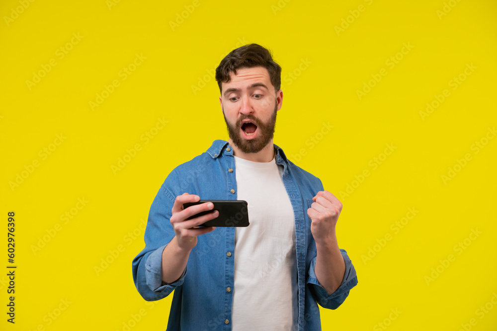 Overjoyed happy tanned handsome man in blue basic t-shirt raise fist up using phone posing isolated on orange yellow studio background. Copy space Banner Mockup. Online Social media. Lucky win concept Overjoyed happy tanned handsome man in blue basic t-shirt raise fist up using phone posing isolated on orange yellow studio background. Copy space Banner Mockup. Online Social media. Lucky win concept