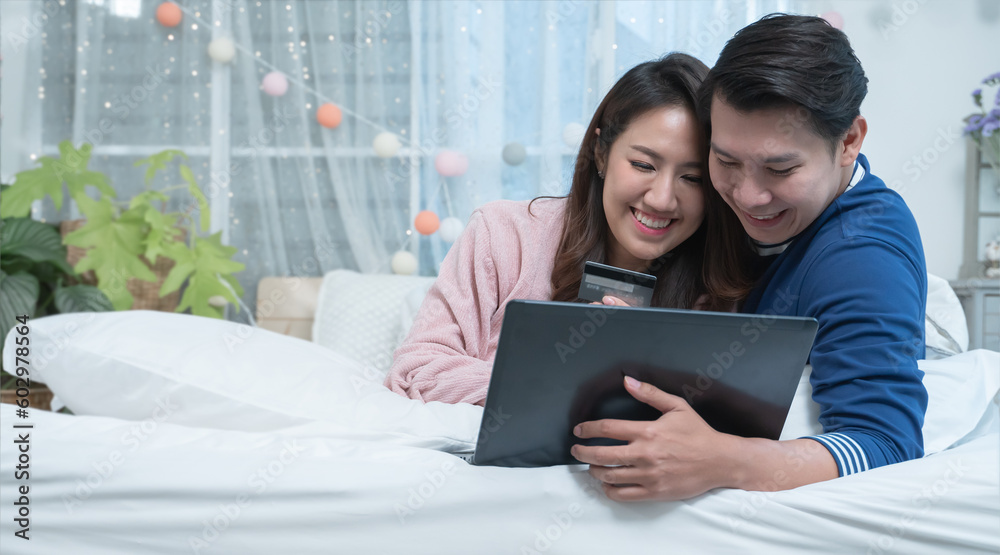 Fototapeta premium Happy asian couple lying on bed, shopping online, using laptop and credit card at home