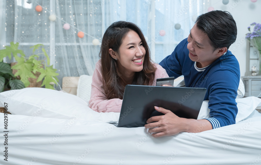 Happy asian couple lying on bed, shopping online, using laptop and credit card at home Happy asian couple lying on bed, shopping online, using laptop and credit card at home