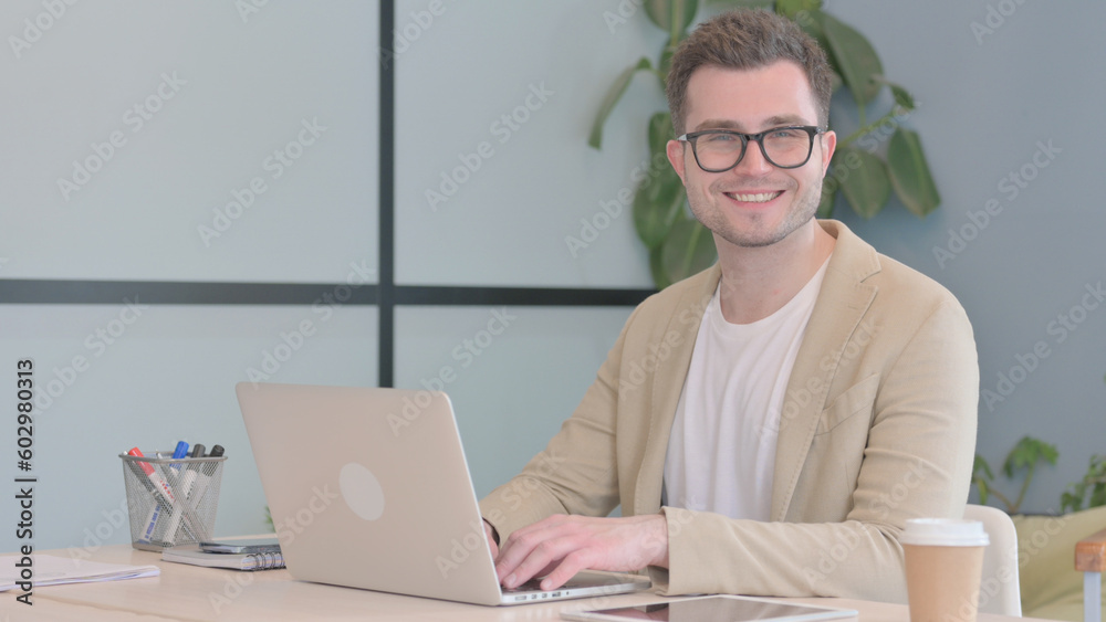 Young Businessman Smiling at Camera while Working on Laptop Young Businessman Smiling at Camera while Working on Laptop