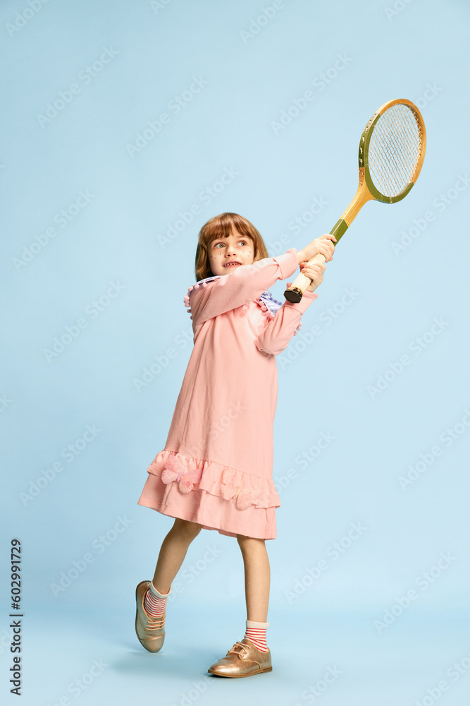 Full-length portrait of little girl in pink dress posing with tennis racket, playing against blue studio background. Concept of childhood, emotions, fun, fashion, active lifestyle, sport Full-length portrait of little girl in pink dress posing with tennis racket, playing against blue studio background. Concept of childhood, emotions, fun, fashion, active lifestyle, sport