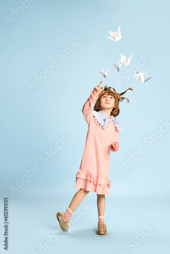 Full-length portrait of playful, lovely little girl in pink dress posing, playing with paper birds against blue studio background. Concept of childhood, emotions, fun, fashion, lifestyle, imagination photo