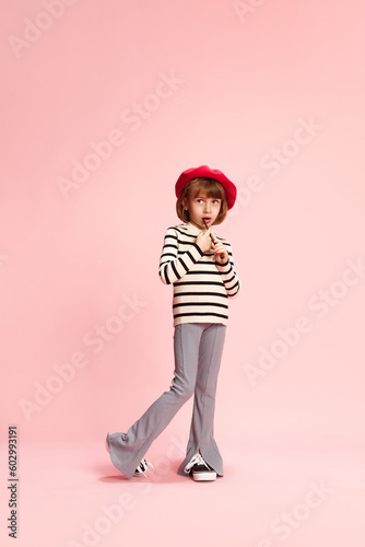 Full-length portrait of little girl, child in red beret and striped sweater posing with painting brush against pink studio background. Concept of childhood, emotions, fun, fashion, lifestyle