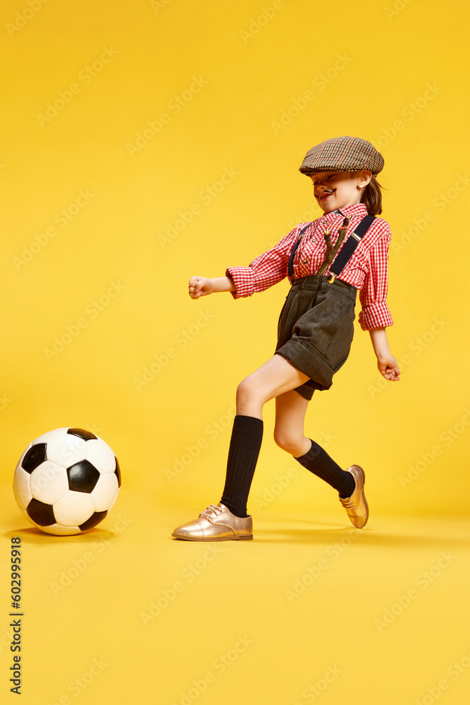 Portrait of stylish little girl, child in retro clothes, cap, shirt and shorts playing with football ball against yellow studio background. Concept of childhood, emotions, fun, fashion, lifestyle Portrait of stylish little girl, child in retro clothes, cap, shirt and shorts playing with football ball against yellow studio background. Concept of childhood, emotions, fun, fashion, lifestyle