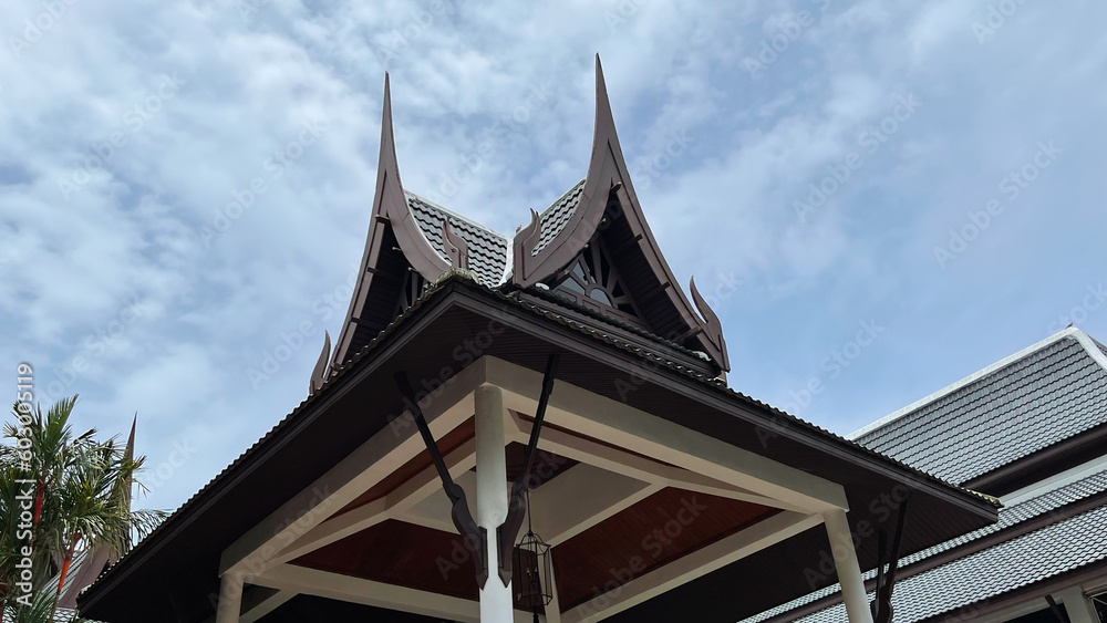 Traditional Thai style roof on a cloudy sky background. Bottom up view. Wooden stylish structure. Top of the gazebo in the tropical garden. Asian architecture. Horizontal frame, close-up photo.