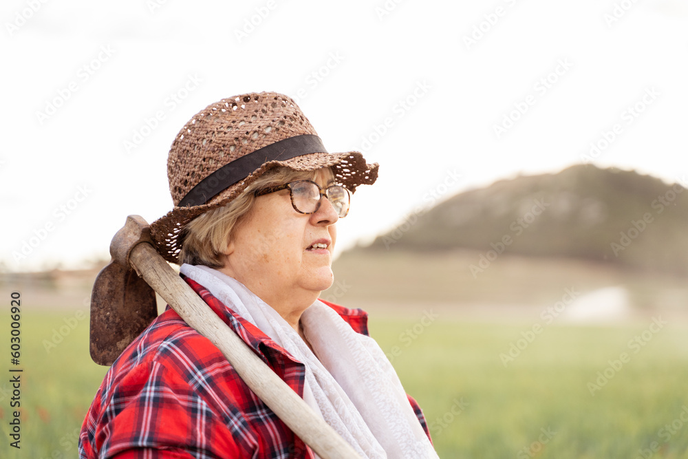 portrait of a grandmother of Caucasian origin wearing a straw hat and red plaid shirt holds her hoe over her shoulder