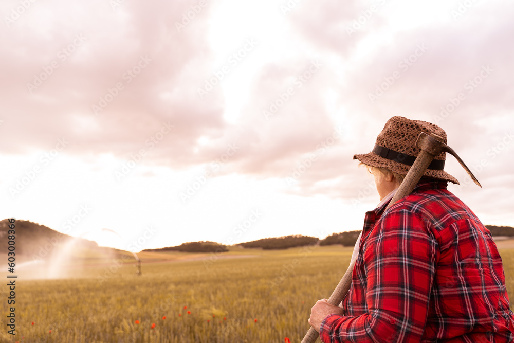Grandmother dressed in straw hat and red plaid shirt holds her hoe and watches water irrigating crop . Photos of people farming in the field.