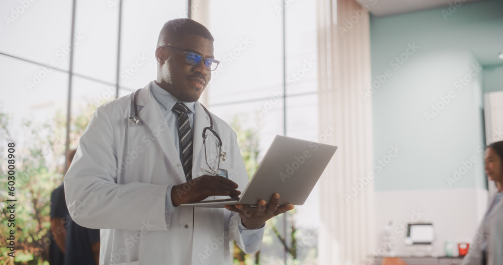 Young African Male Doctor Wearing White Coat, Using a Laptop Computer while Standing in Hospital Hallway. Handsome Black Medical Health Care Professional Working with Patient Test Results Young African Male Doctor Wearing White Coat, Using a Laptop Computer while Standing in Hospital Hallway. Handsome Black Medical Health Care Professional Working with Patient Test Results