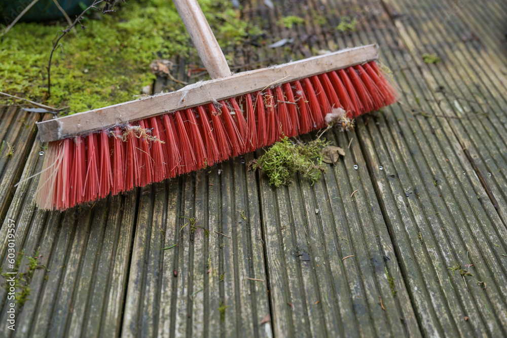 Outdoor broom with red plastic bristles on a weathered wooden deck with slippery algae and moss, spring cleaning in garden and yard, copy space, selected focus