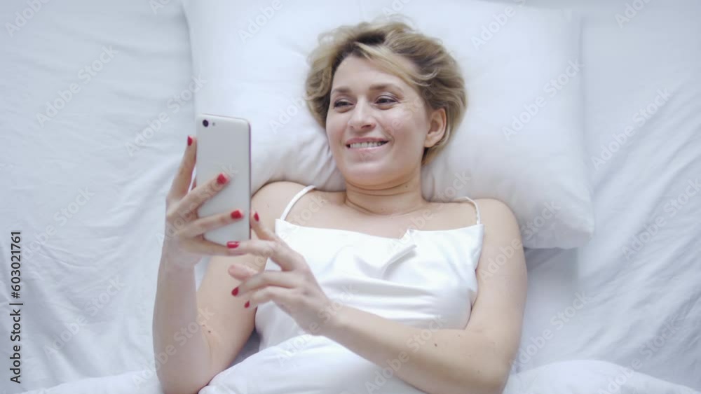 Happy satisfied woman lying in her bed and smiling, booking vacations tour or tickets on favourite music band online via smartphone, doing shopping online Happy satisfied woman lying in her bed and smiling, booking vacations tour or tickets on favourite music band online via smartphone, doing shopping online