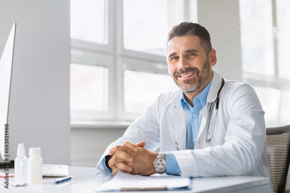 Portrait of friendly middle aged male doctor wearing medical workwear posing at clinic, sitting at workdesk