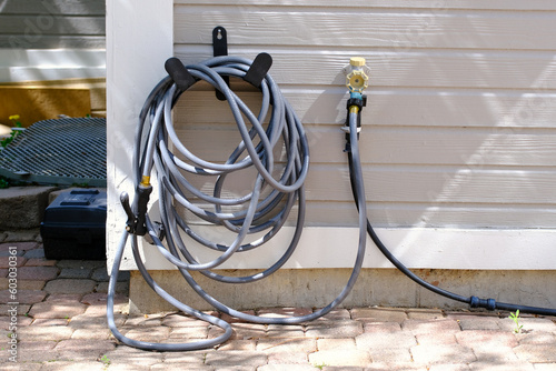 Garden hose with a sprayer connected to house tap water supply, coiled, hanging on a wall of a residential home building in the backyard patio. Gardening appliances. Irrigation equipment. Home chores.