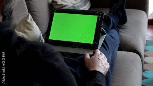 Man sitting on sofa at home holding green screen tablet. Watching entertainment content or learning.
