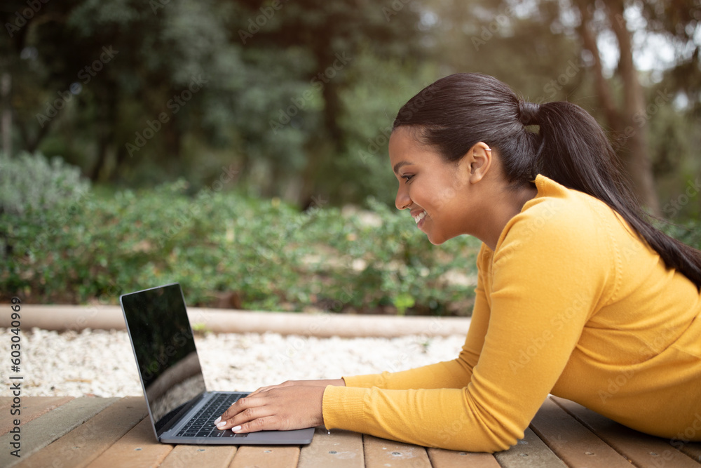 Happy brazilian college student girl preparing for exams or doing homework on laptop, lying on bench in park, side view Happy brazilian college student girl preparing for exams or doing homework on laptop, lying on bench in park, side view