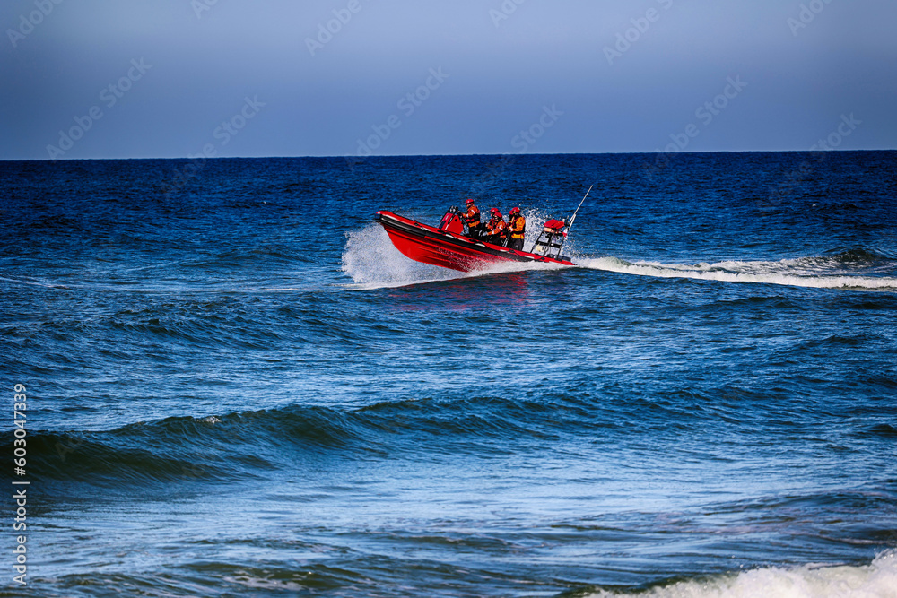 Pomarańczowa łódka z ratownikami płynie z akcją ratowniczą na morzu.  - obrazy, fototapety, plakaty 