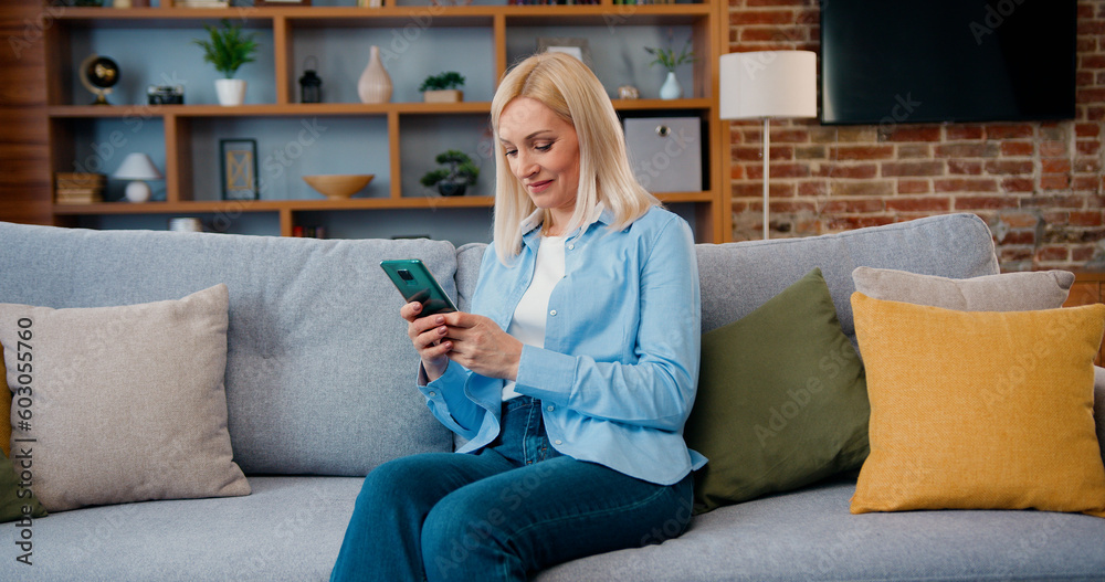 Smiling middle aged lady customer texting mobile sms message playing mobile games using social media apps at home. Millennial woman spending time at home with cell gadget technology. Smiling middle aged lady customer texting mobile sms message playing mobile games using social media apps at home. Millennial woman spending time at home with cell gadget technology.